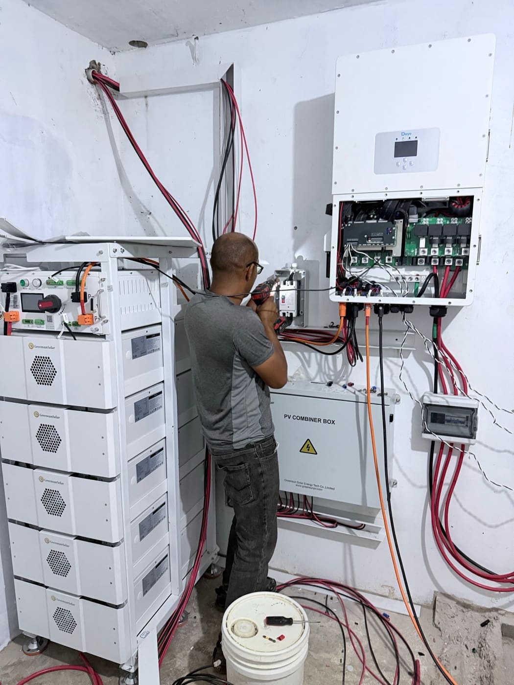 A technician working inside an open hybrid inverter beside a stacked battery rack